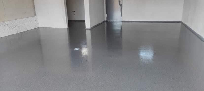 Szara, błyszcząca podłoga w pustym pomieszczeniu z białymi ścianami i widocznymi drzwiami w tle. Podłoga ma delikatną strukturę i odbija światło.