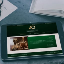 Kancelaria Adwokacka Adwokat Aleksandra Dzida - Tablet wyświetlający stronę internetową kancelarii adwokackiej z logiem, adresem i informacjami o adwokacie Aleksandrze Dzidzie, obok leżą okulary, długopis i notatnik na ciemnym tle.