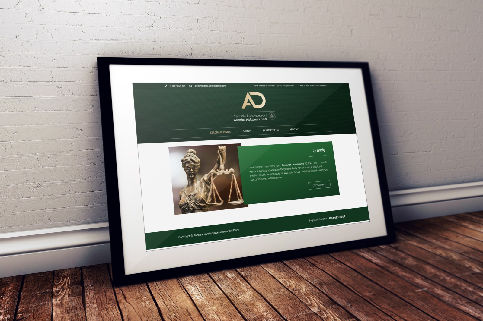 Zdjęcie strony internetowej kancelarii adwokackiej Aleksandra Dzida, z brązową statuetką Temidy w sekcji 'O mnie', umieszczone w ramce na drewnianej podłodze i przy białej ceglanej ścianie.