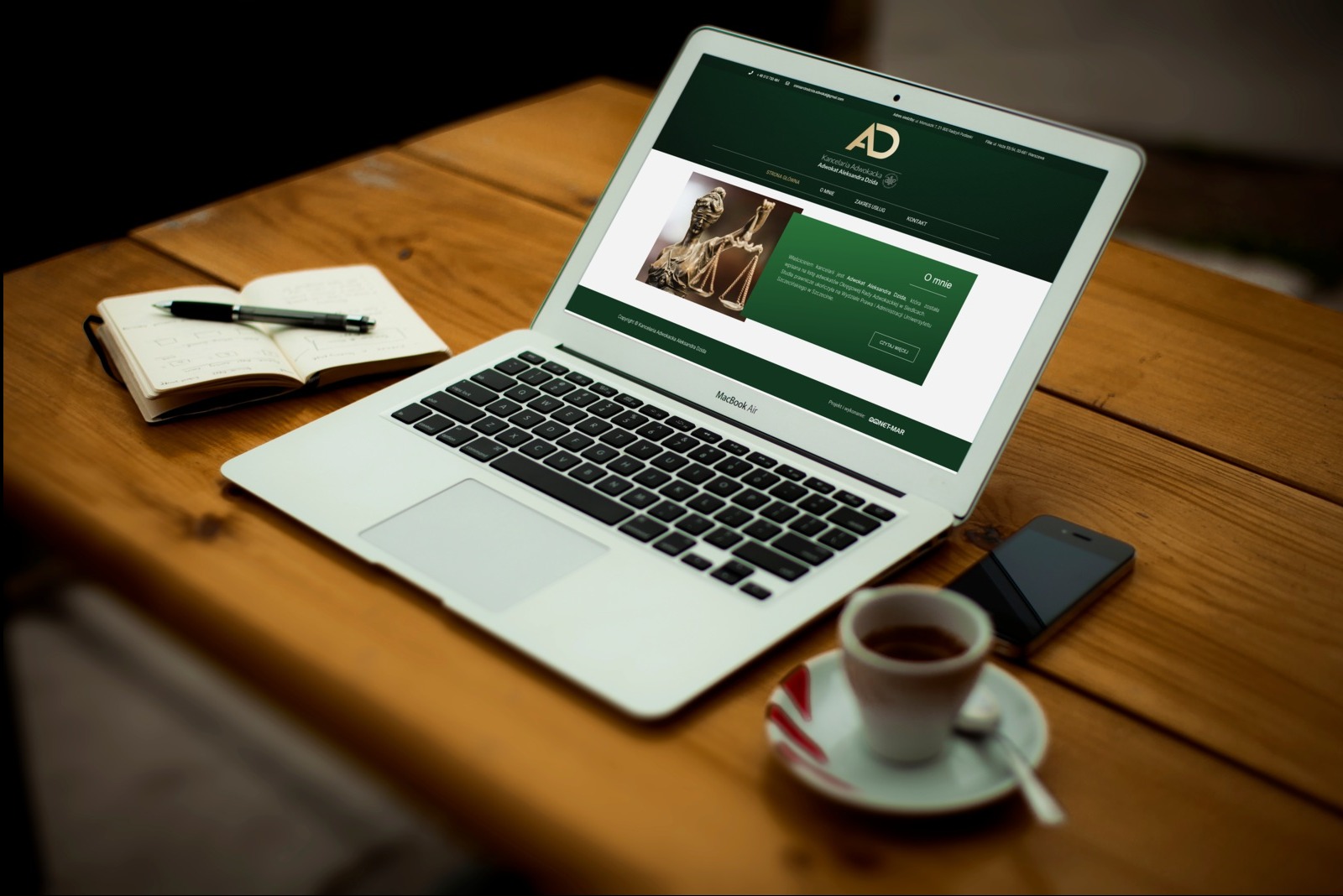 Laptop z otwartą stroną internetową kancelarii adwokackiej na drewnianym stole, obok notatnik z długopisem, smartfon i filiżanka kawy.