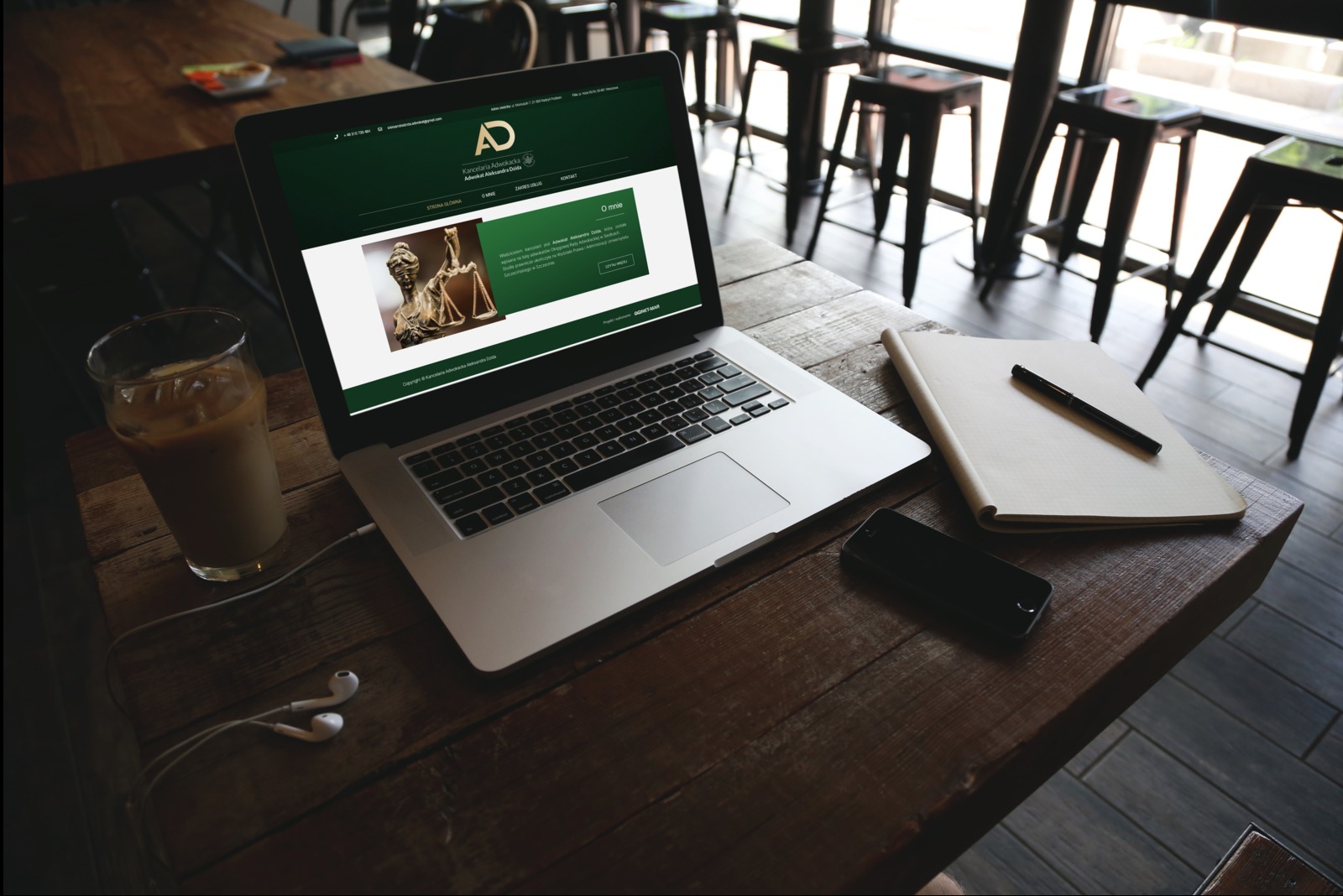 Laptop wyświetlający stronę internetową kancelarii prawnej z symbolem Temidy, obok szklanka z kawą, notatnik, długopis, telefon i słuchawki na drewnianym biurku.