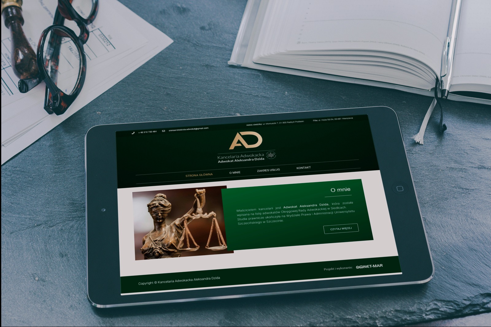 Tablet wyświetlający stronę internetową kancelarii adwokackiej z logiem, adresem i informacjami o adwokacie Aleksandrze Dzidzie, obok leżą okulary, długopis i notatnik na ciemnym tle.