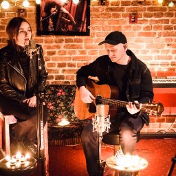 LOT ĆMY - Pracownia muzyczna - Duet muzyczny w kameralnym studio: wokalistka przy mikrofonie, gitarzysta grający na gitarze akustycznej, w tle ceglana ściana, keyboard i świecące lampki.