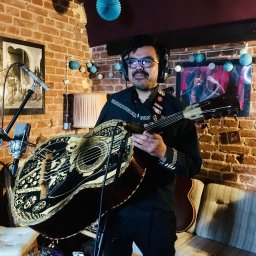 LOT ĆMY - Pracownia muzyczna - Muzyk z wąsami w słuchawkach, trzymający ozdobną gitarę bajo sexto w ceglanym pokoju z mikrofonem i dekoracjami.