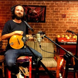 LOT ĆMY - Pracownia muzyczna - Młody mężczyzna z brodą i słuchawkami, siedzący na stołku w studio nagrań, trzymający mandolinę przed mikrofonem, z ceglaną ścianą i klawiszami w tle.