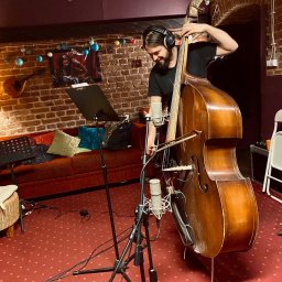 LOT ĆMY - Pracownia muzyczna - Młody muzyk z brodą i słuchawkami gra na kontrabasie w kameralnym studiu nagrań, z mikrofonami i nutami w tle, czerwona wykładzina z wzorem na podłodze.