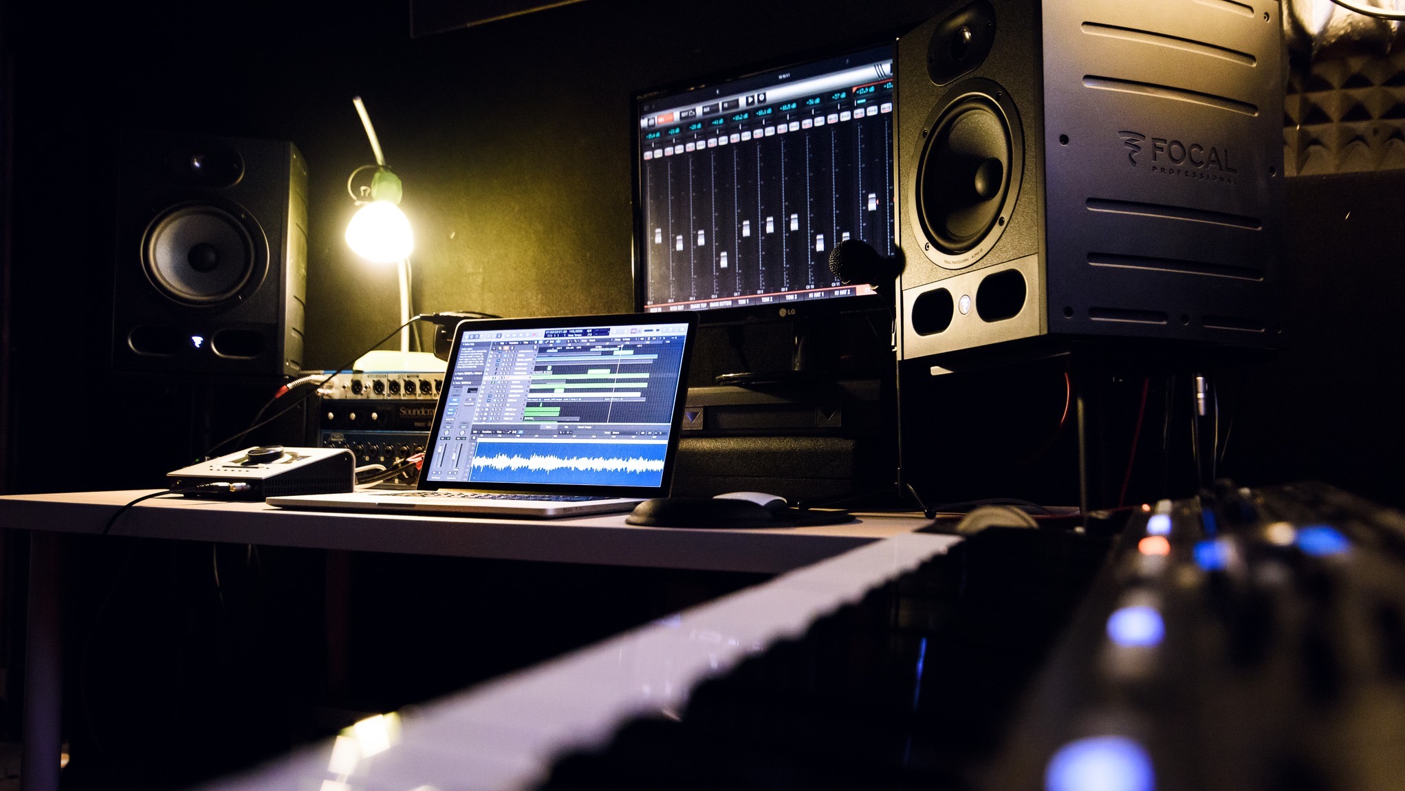 Profesjonalne studio nagrań z laptopem wyświetlającym program do edycji audio, monitory studyjne Focal, klawiatura MIDI na pierwszym planie i lampka biurkowa oświetlająca sprzęt.
