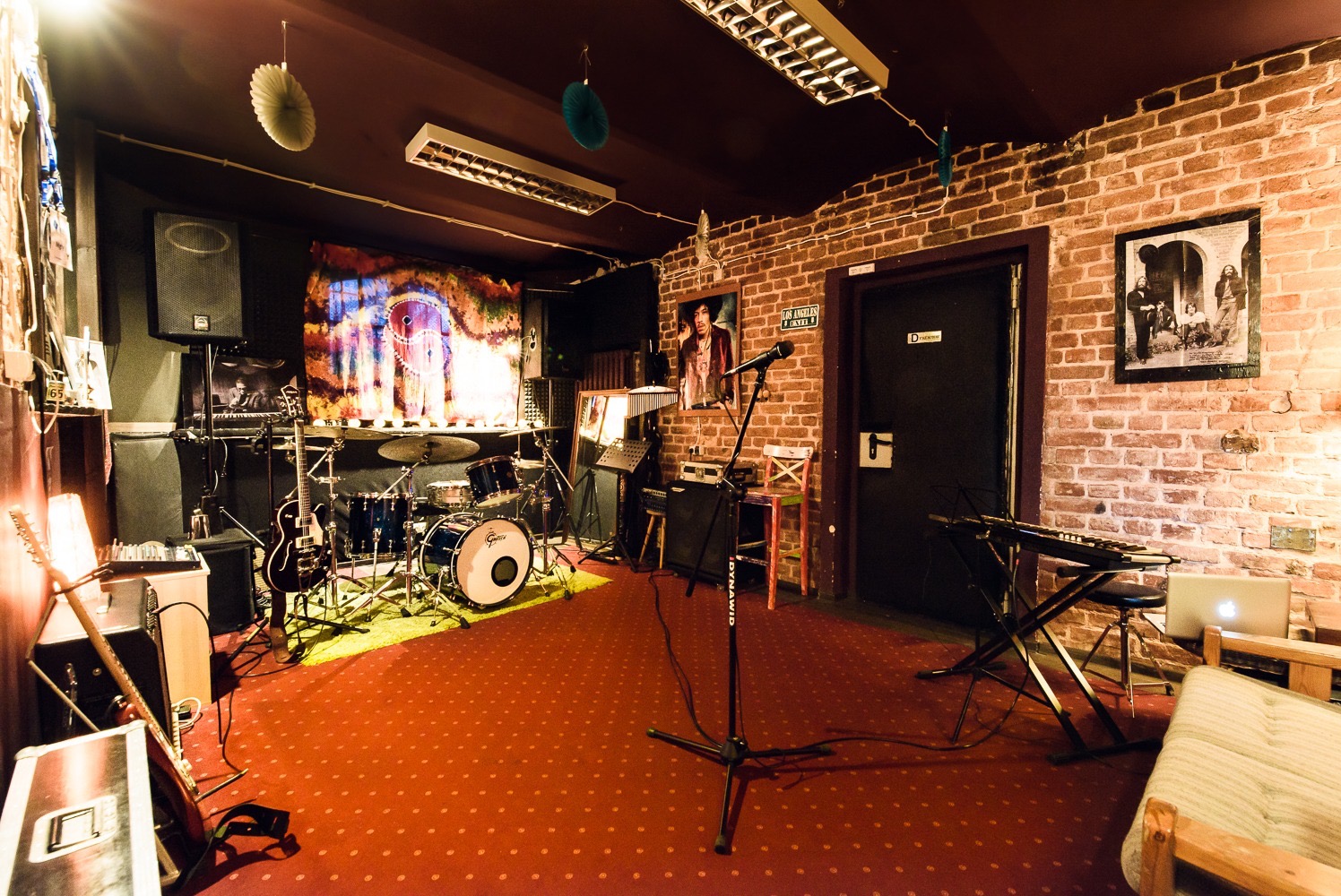 Wnętrze studia nagraniowego z instrumentami: perkusja, gitara, keyboard, mikrofon na statywie, wzmacniacze, czerwona wykładzina, ceglana ściana z plakatami.