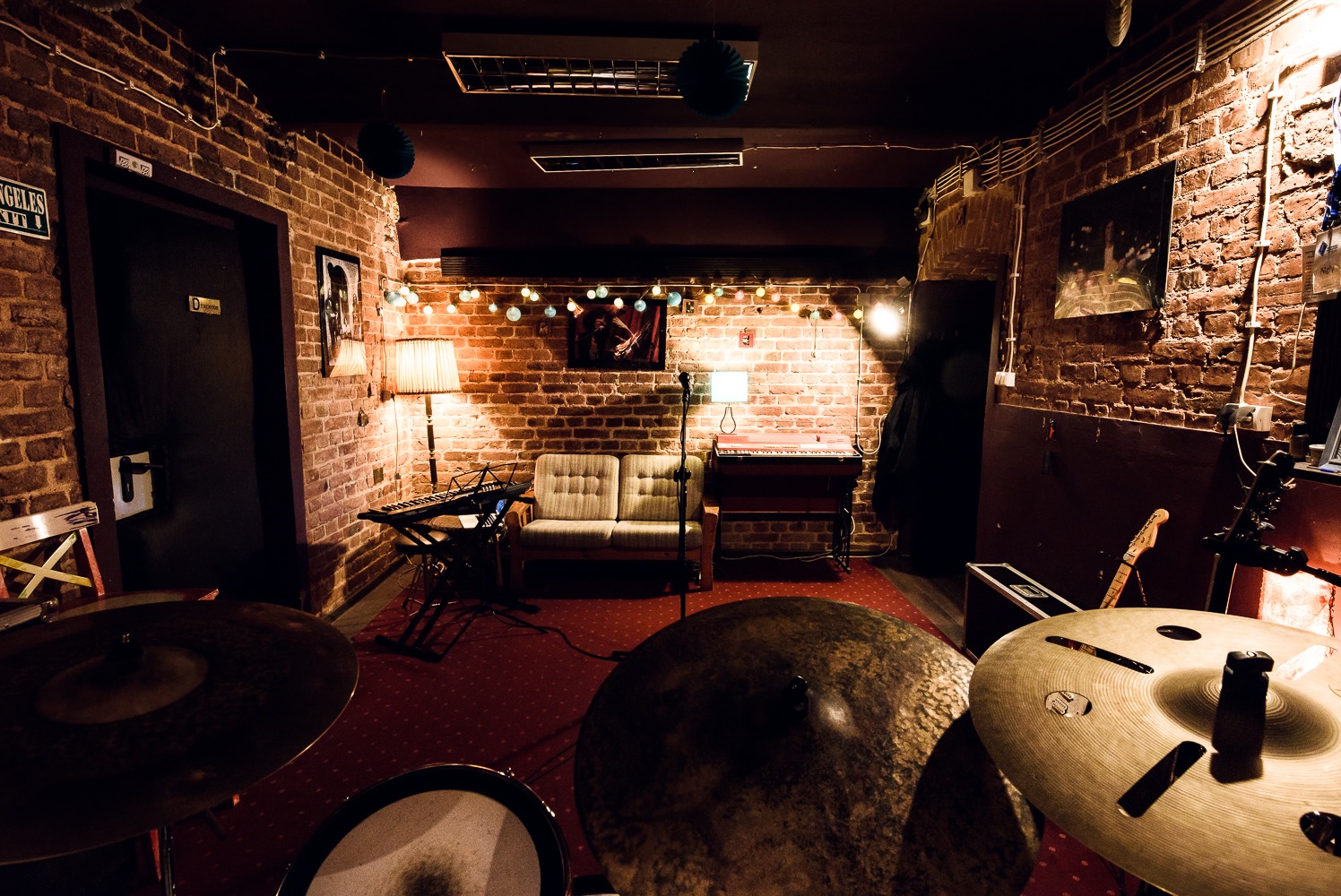 Wnętrze studia nagrań z surową cegłą, oświetlone girlandami żarówek, z widocznymi instrumentami perkusyjnymi na pierwszym planie i sofą w tle.
