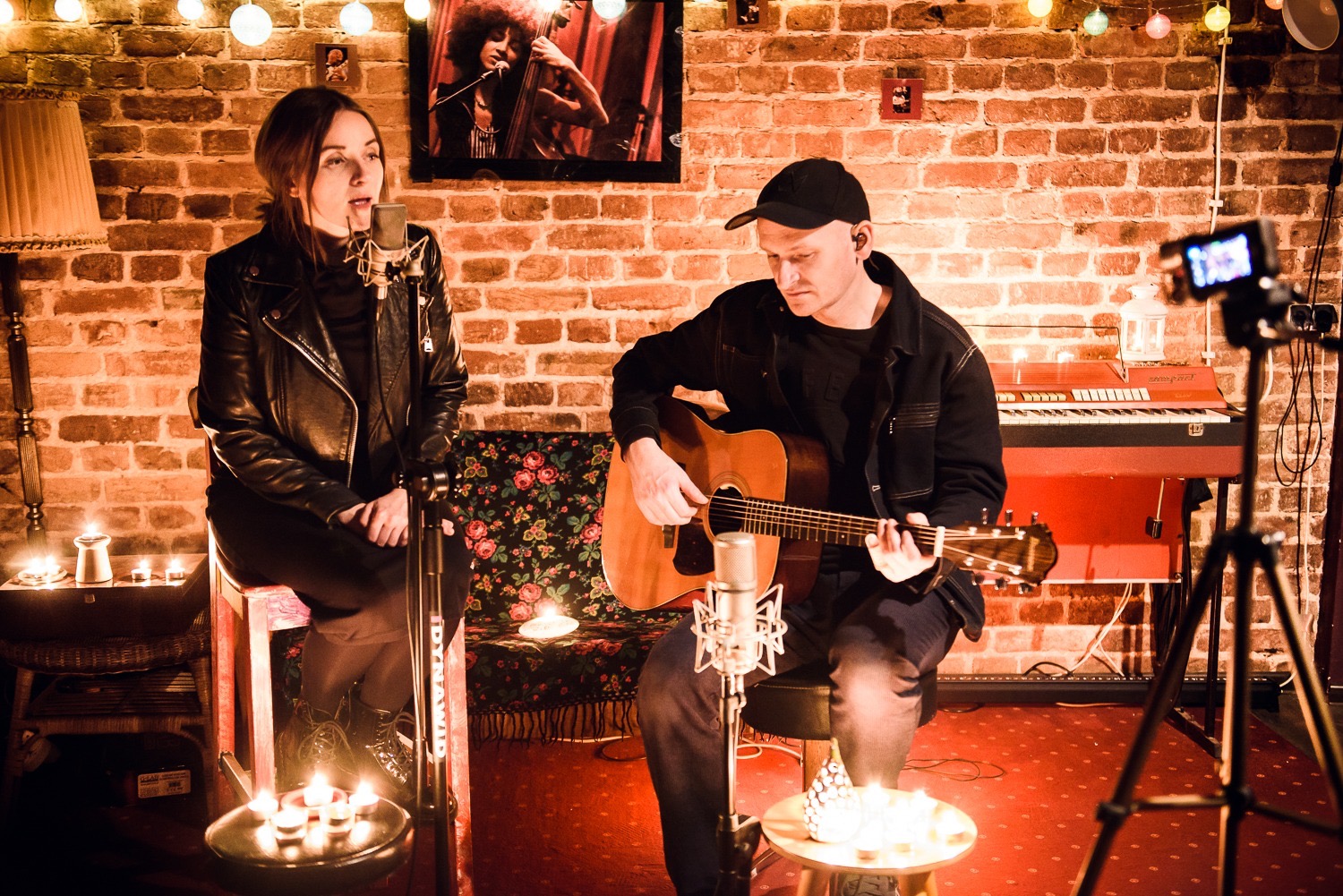 Duet muzyczny w kameralnym studio: wokalistka przy mikrofonie, gitarzysta grający na gitarze akustycznej, w tle ceglana ściana, keyboard i świecące lampki.