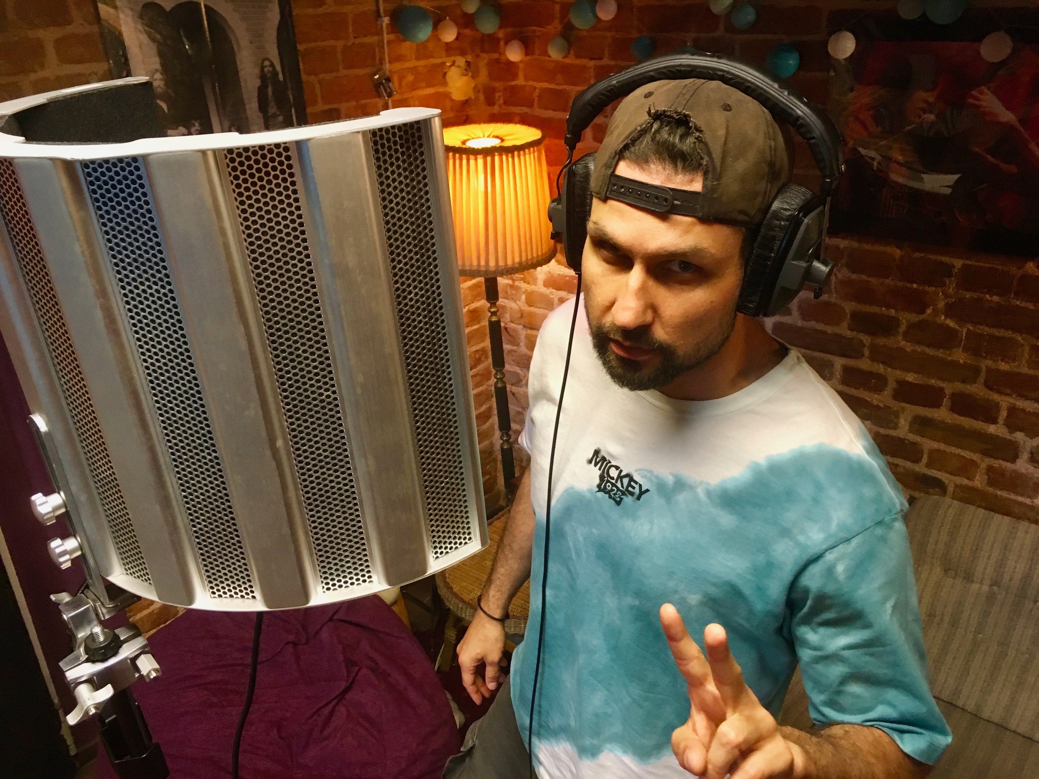 Mężczyzna w słuchawkach studyjnych pozuje w domowym studiu nagrań z ekranem akustycznym, ceglaną ścianą i lampką w tle, pokazując znak zwycięstwa.