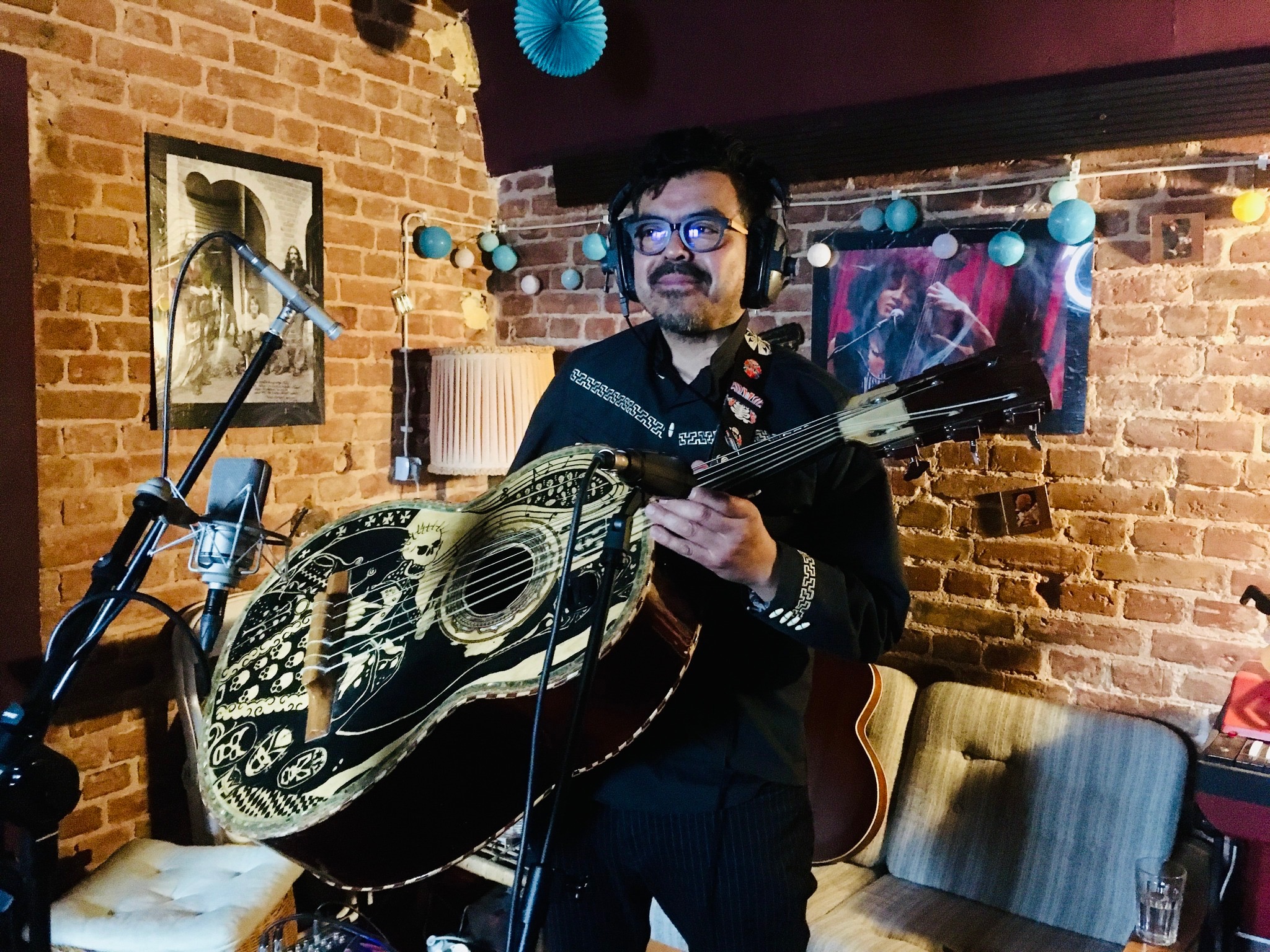 Muzyk z wąsami w słuchawkach, trzymający ozdobną gitarę bajo sexto w ceglanym pokoju z mikrofonem i dekoracjami.