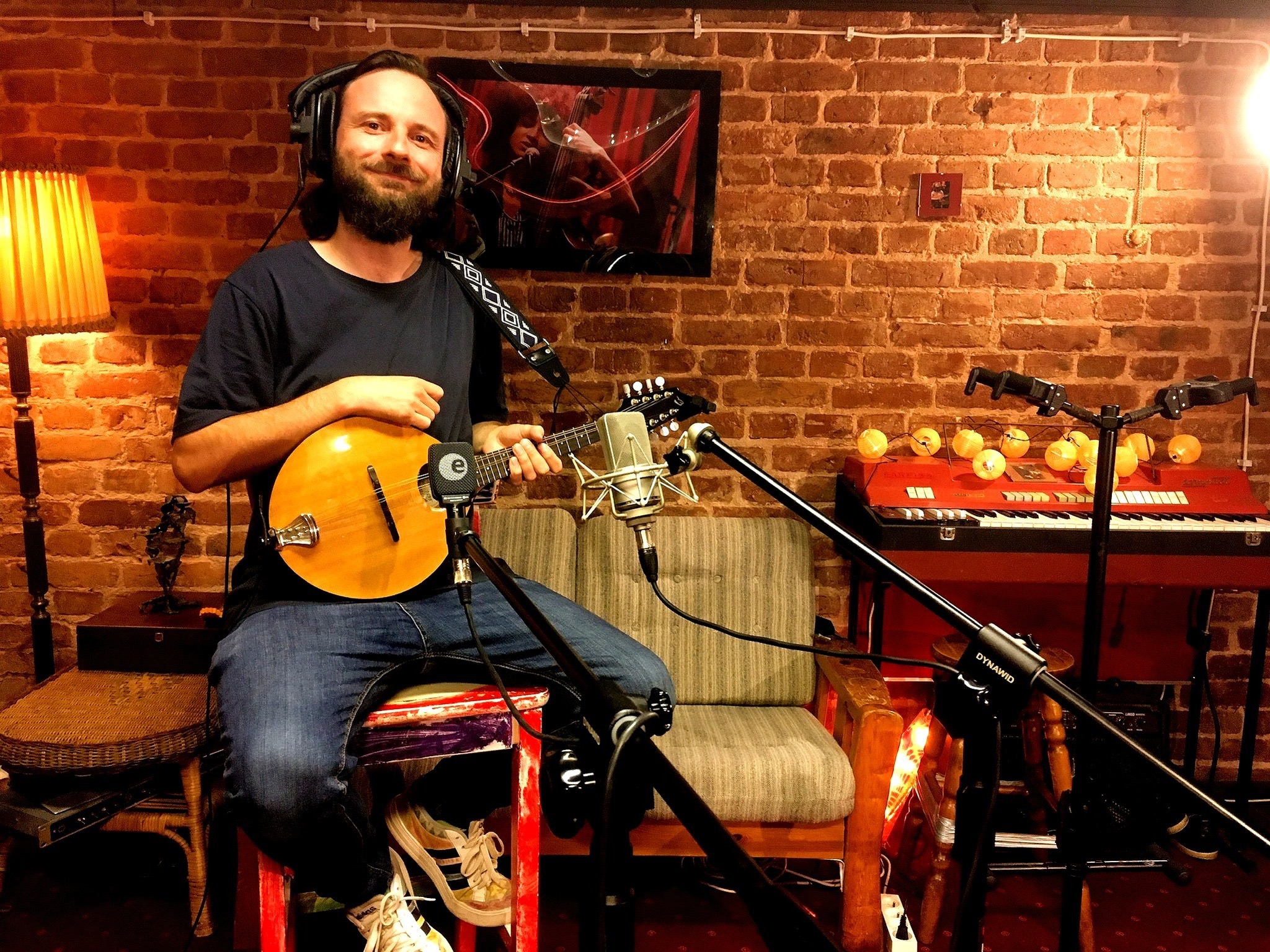 Młody mężczyzna z brodą i słuchawkami, siedzący na stołku w studio nagrań, trzymający mandolinę przed mikrofonem, z ceglaną ścianą i klawiszami w tle.