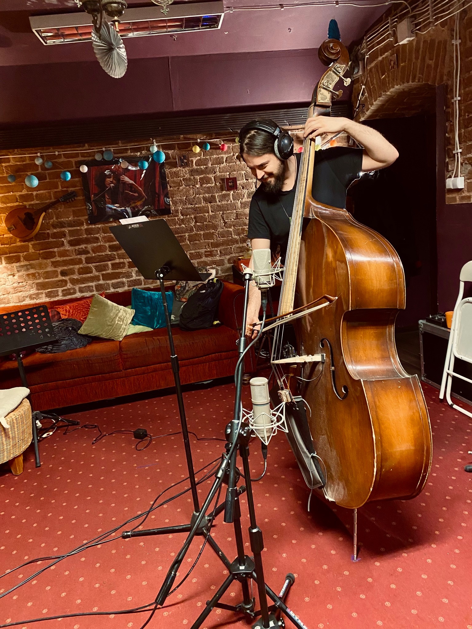Młody muzyk z brodą i słuchawkami gra na kontrabasie w kameralnym studiu nagrań, z mikrofonami i nutami w tle, czerwona wykładzina z wzorem na podłodze.