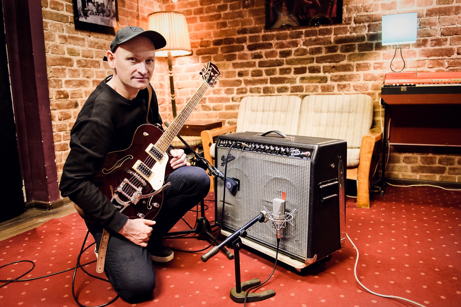 Gitarzysta w czarnej bluzie i czapce trzymający bordową gitarę elektryczną typu hollow body, obok wzmacniacza gitarowego i mikrofonu w pomieszczeniu z ceglaną ścianą i czerwonym dywanem.