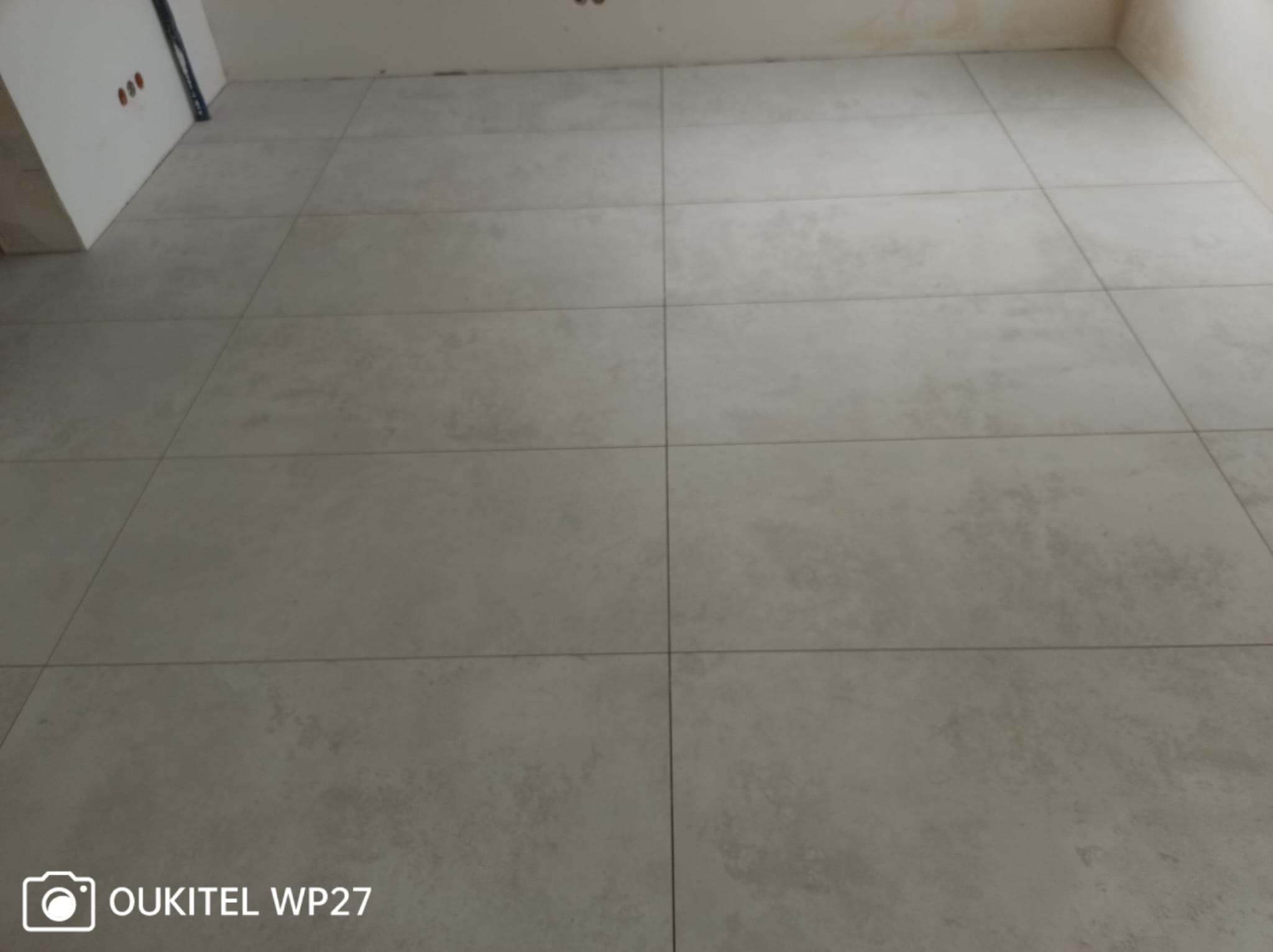 Ułożona podłoga z szarych, matowych płytek ceramicznych o dużym formacie, widoczne fugi i fragment ściany z otworami na gniazdka.
