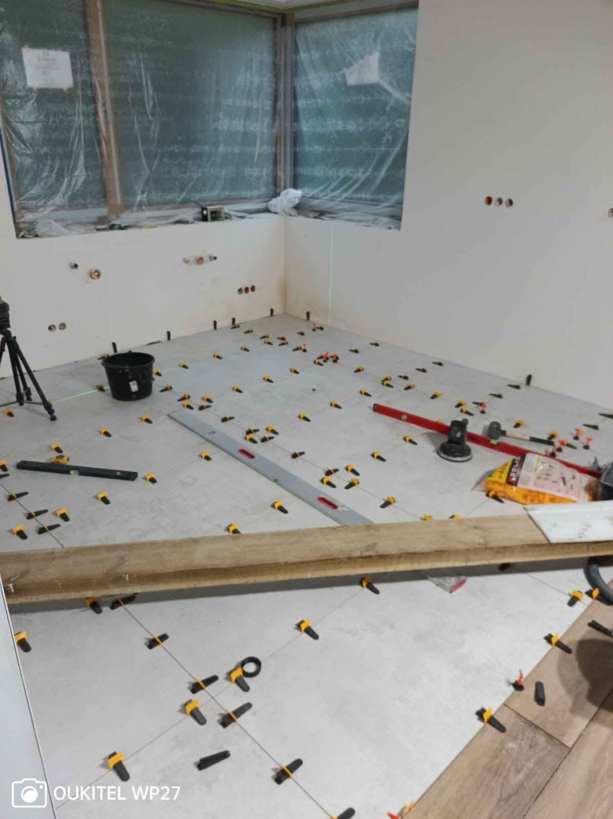 Podłoga w trakcie układania płytek, widoczne poziomice, krzyżyki dystansowe, laser krzyżowy, okno zabezpieczone folią.