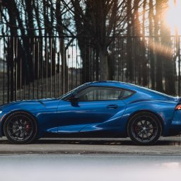 Krysik Studio - Niebieska Toyota Supra na tle metalowego ogrodzenia i drzew, z promieniami słońca prześwitującymi przez gałęzie.