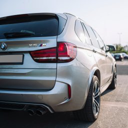 Krysik Studio - Srebrny samochód BMW X5M z widocznymi tylnymi światłami, logiem marki i czterema rurami wydechowymi, sfotografowany na parkingu w słoneczny dzień.