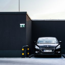 Krysik Studio - Czarny samochód Peugeot zaparkowany przy ciemnym budynku z oznakowaniem drogi ewakuacyjnej i słupkami ochronnymi z żółtymi pasami.