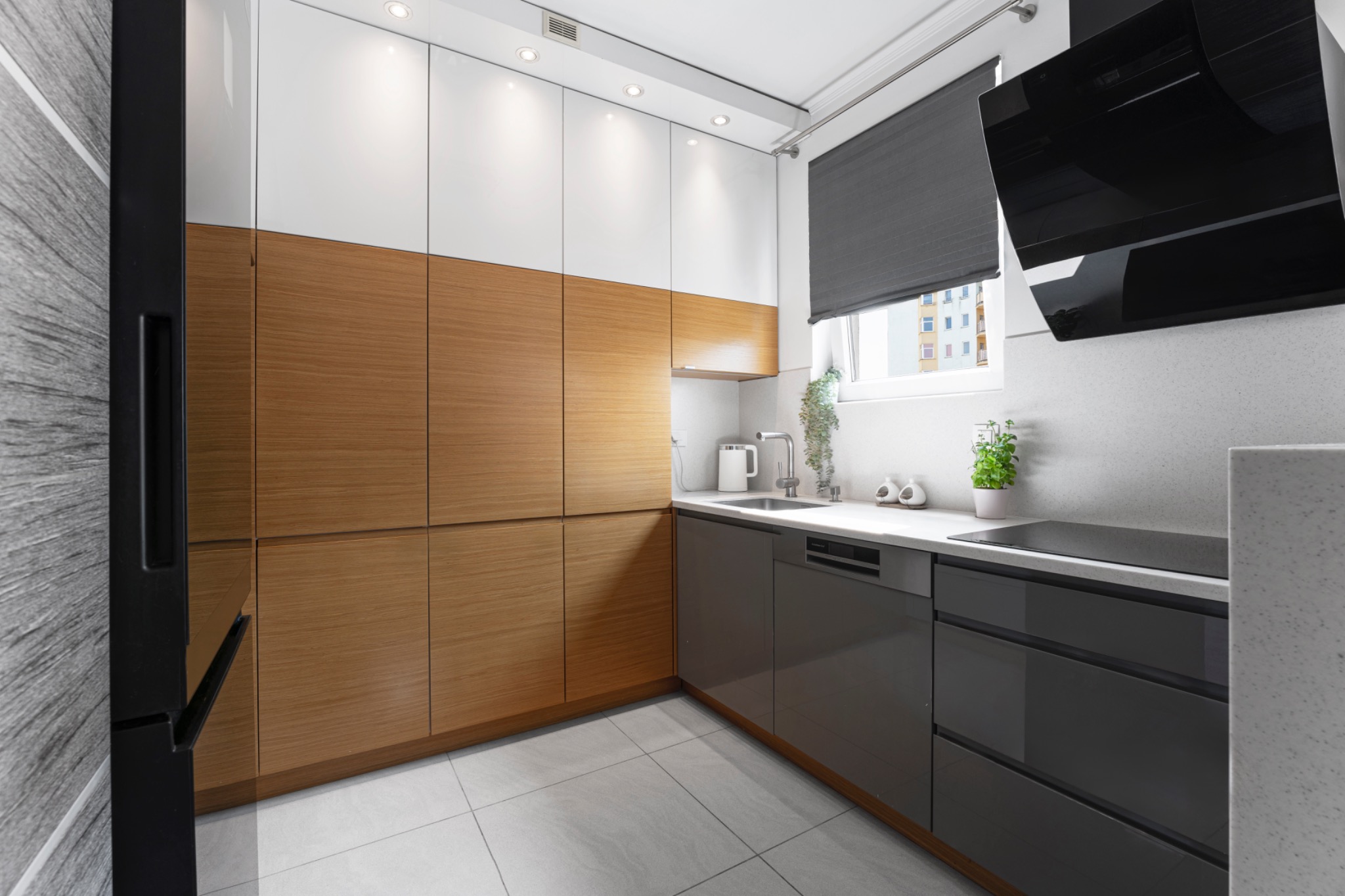Nowoczesna kuchnia z drewnianymi i białymi szafkami, szarymi dolnymi szafkami, czarnym okapem i rośliną w doniczce na blacie.