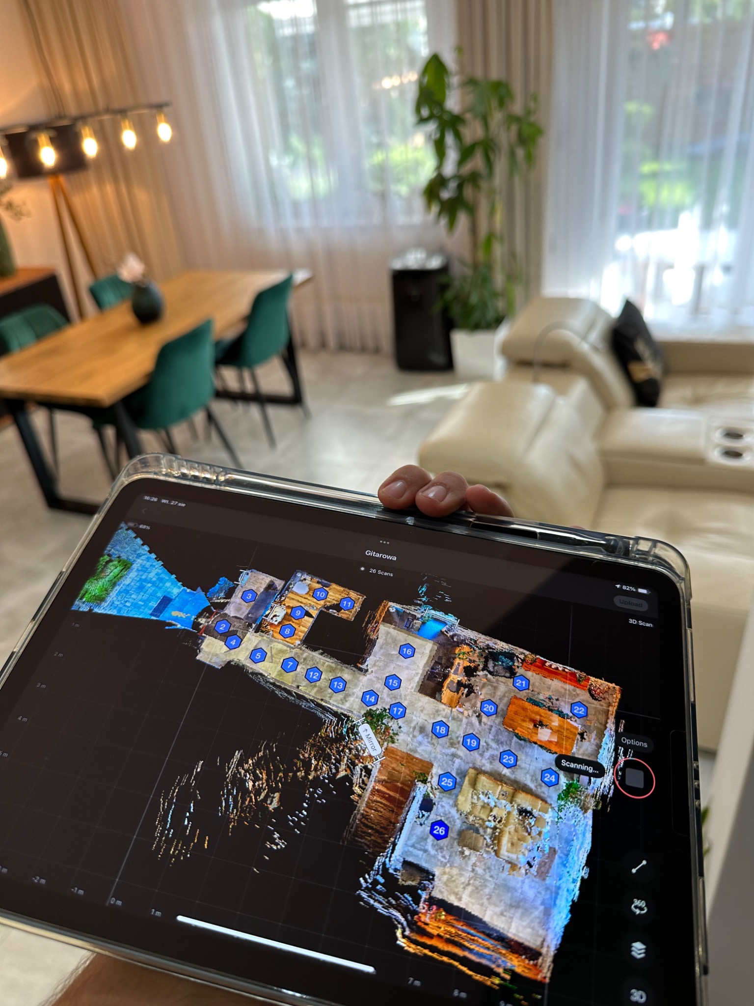 Tablet wyświetla trójwymiarowy plan wnętrza salonu z numerowanymi punktami skanowania; w tle widoczny stół z zielonymi krzesłami, biała sofa i roślina doniczkowa.