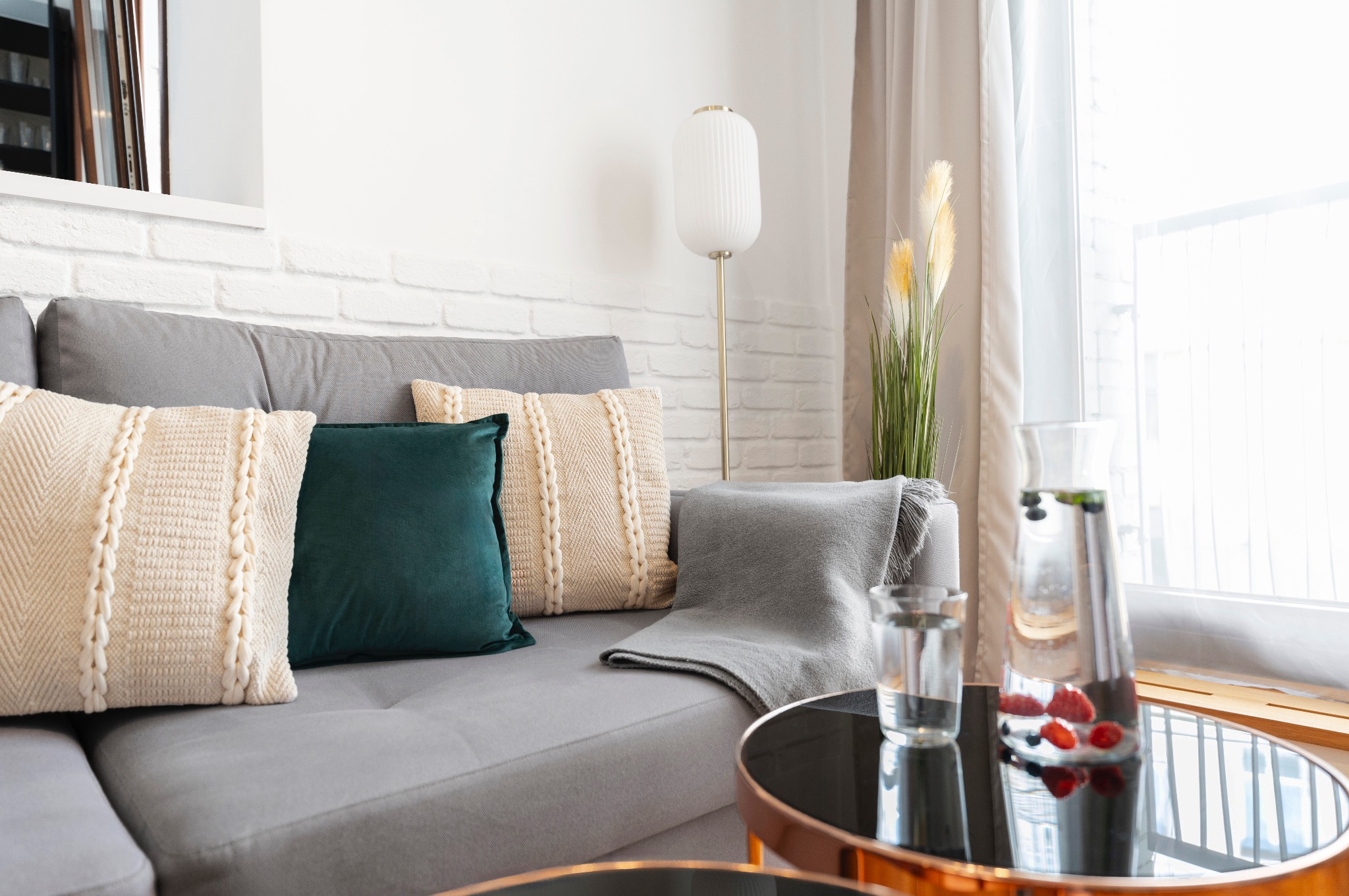 Jasny salon z szarą sofą, dekoracyjnymi poduszkami, lampą podłogową i stolikiem kawowym z napojem owocowym w karafce.
