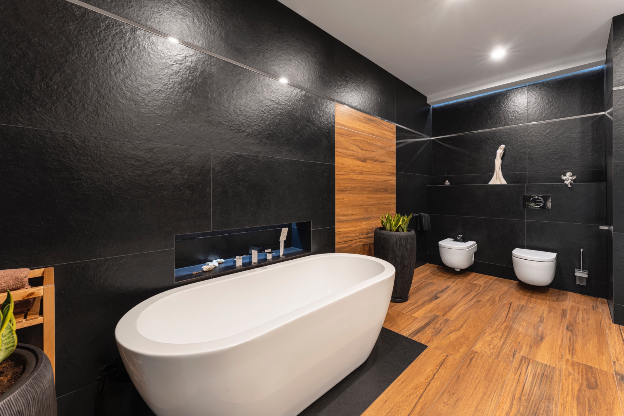 Nowoczesna łazienka z czarnymi płytkami, drewnianą podłogą i wolnostojącą białą wanną, akcentowana rośliną w donicy i subtelnym oświetleniem LED.