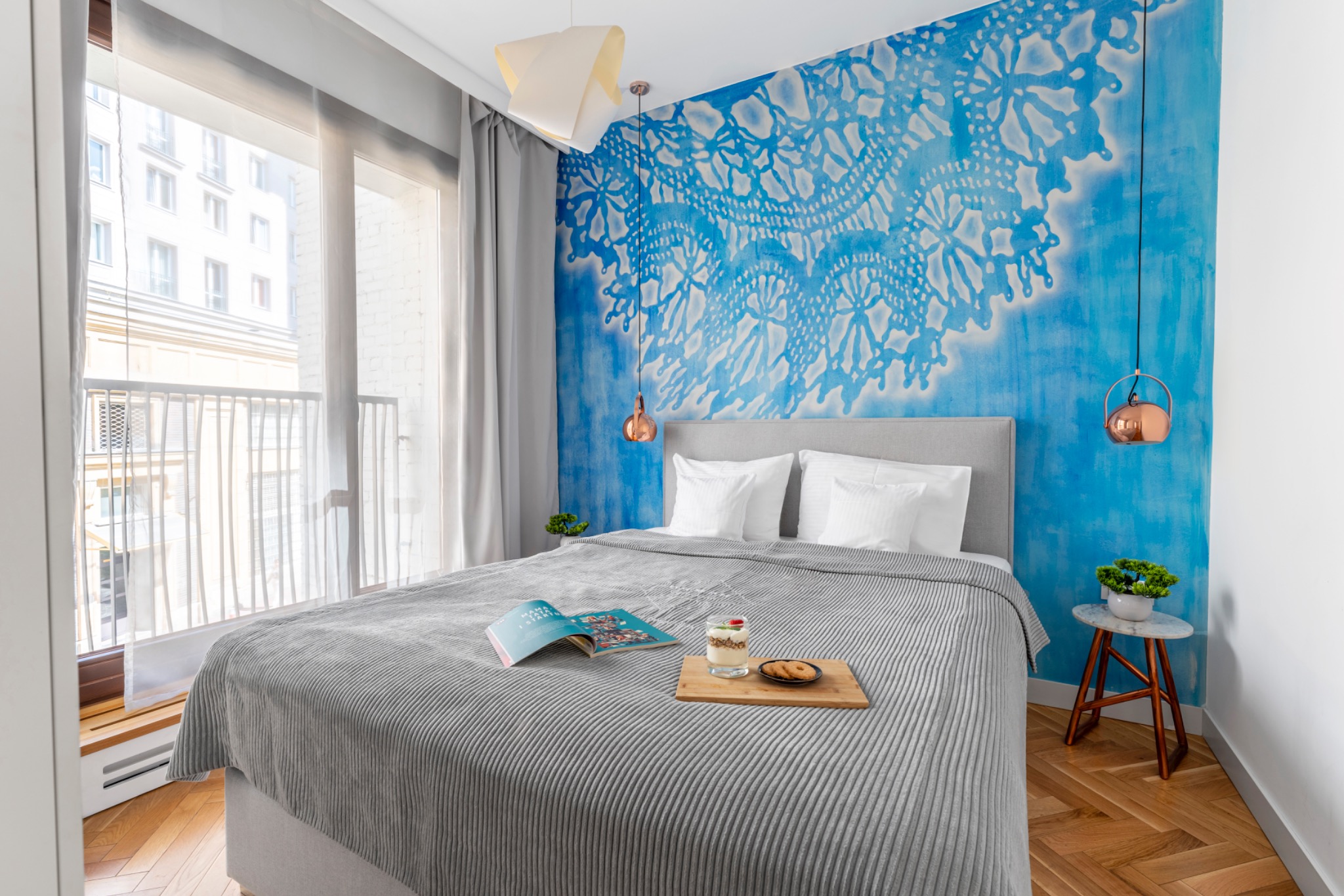 Jasna sypialnia z łóżkiem z szarą narzutą w paski, drewnianą tacą z deserem i otwartą książką, błękitną ścianą z motywem koronki, balkonem i parkietem w jodełkę.