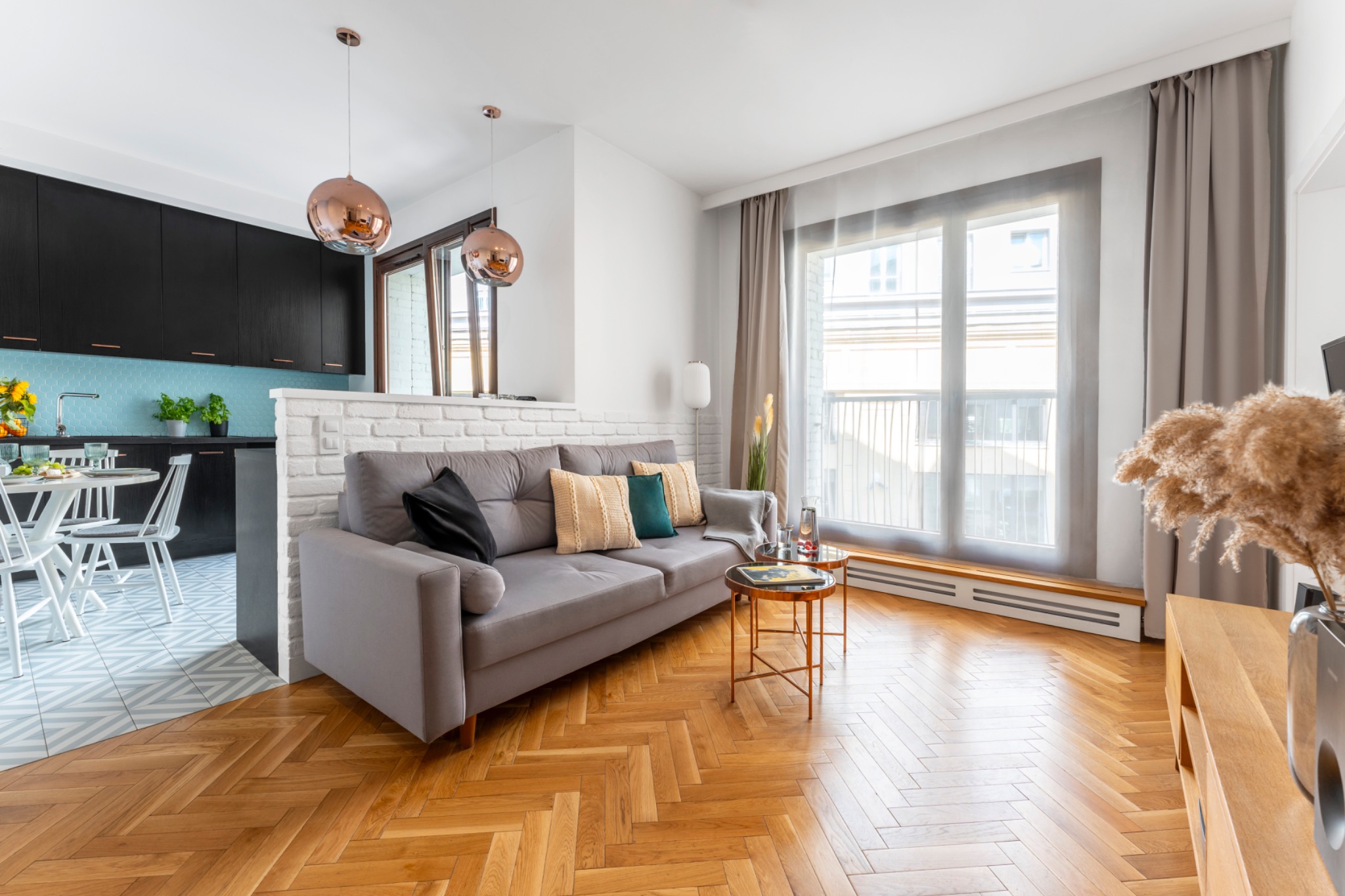 Jasne wnętrze z parkietem w jodełkę, szarą sofą z poduszkami w kolorach beżu, zieleni i czerni, okrągłym stolikiem kawowym i widokiem na kuchnię z czarnymi szafkami i turkusowymi płytkami.