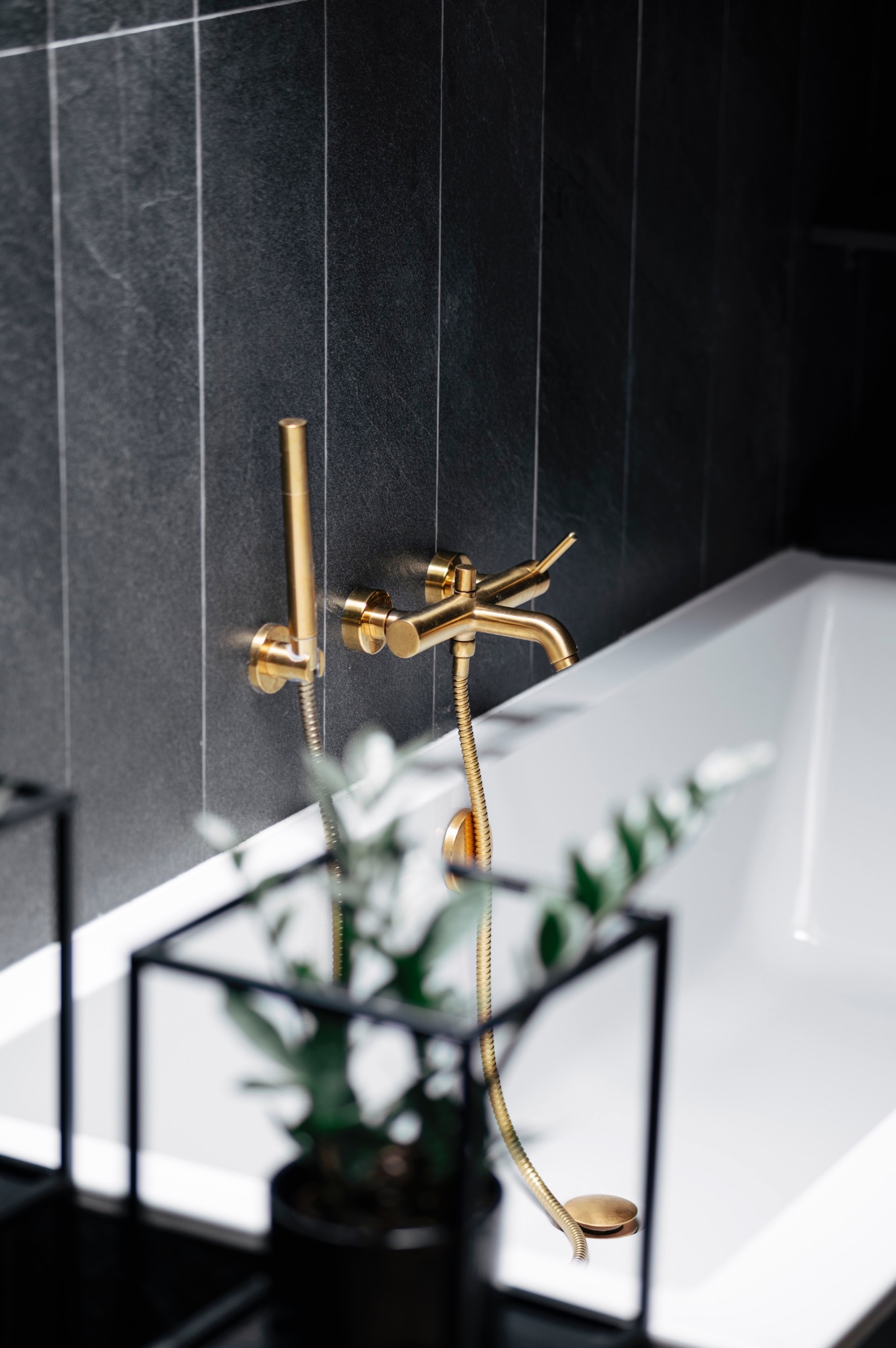 Bateria wannowa w kolorze złotym na tle czarnych, pionowych płytek w łazience, z fragmentem wanny i rośliną doniczkową w kadrze.