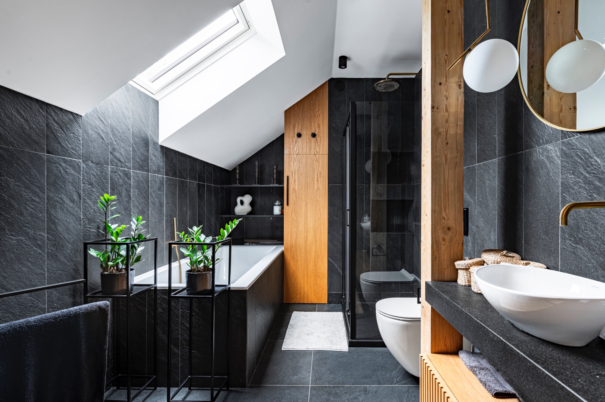 Nowoczesna łazienka na poddaszu z czarnymi płytkami imitującymi łupek, drewnianymi akcentami, owalną umywalką nablatową i roślinami w minimalistycznych donicach.