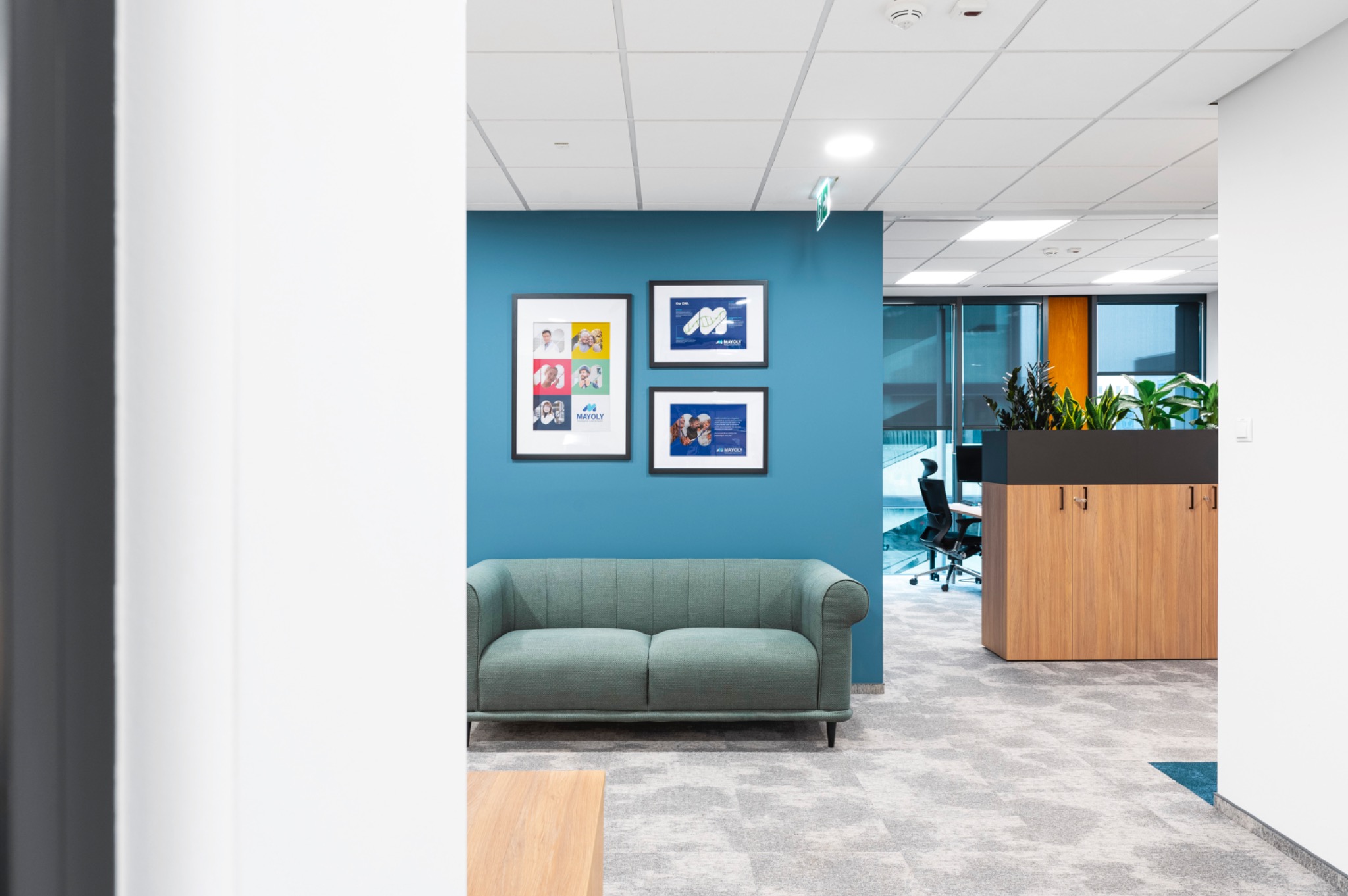 Nowoczesne biuro z turkusową ścianą, zieloną sofą i trzema oprawionymi grafikami firmowymi na ścianie, widok przez białe drzwi.