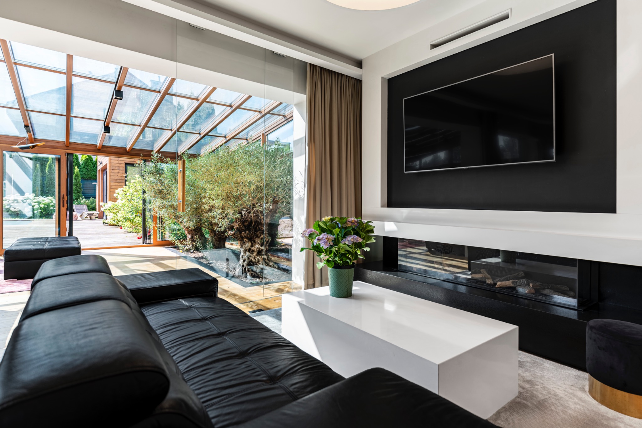 Nowoczesny salon z czarną skórzaną sofą, białym stolikiem kawowym z hortensjami w doniczce i przeszkloną oranżerią wychodzącą na drewniany taras.