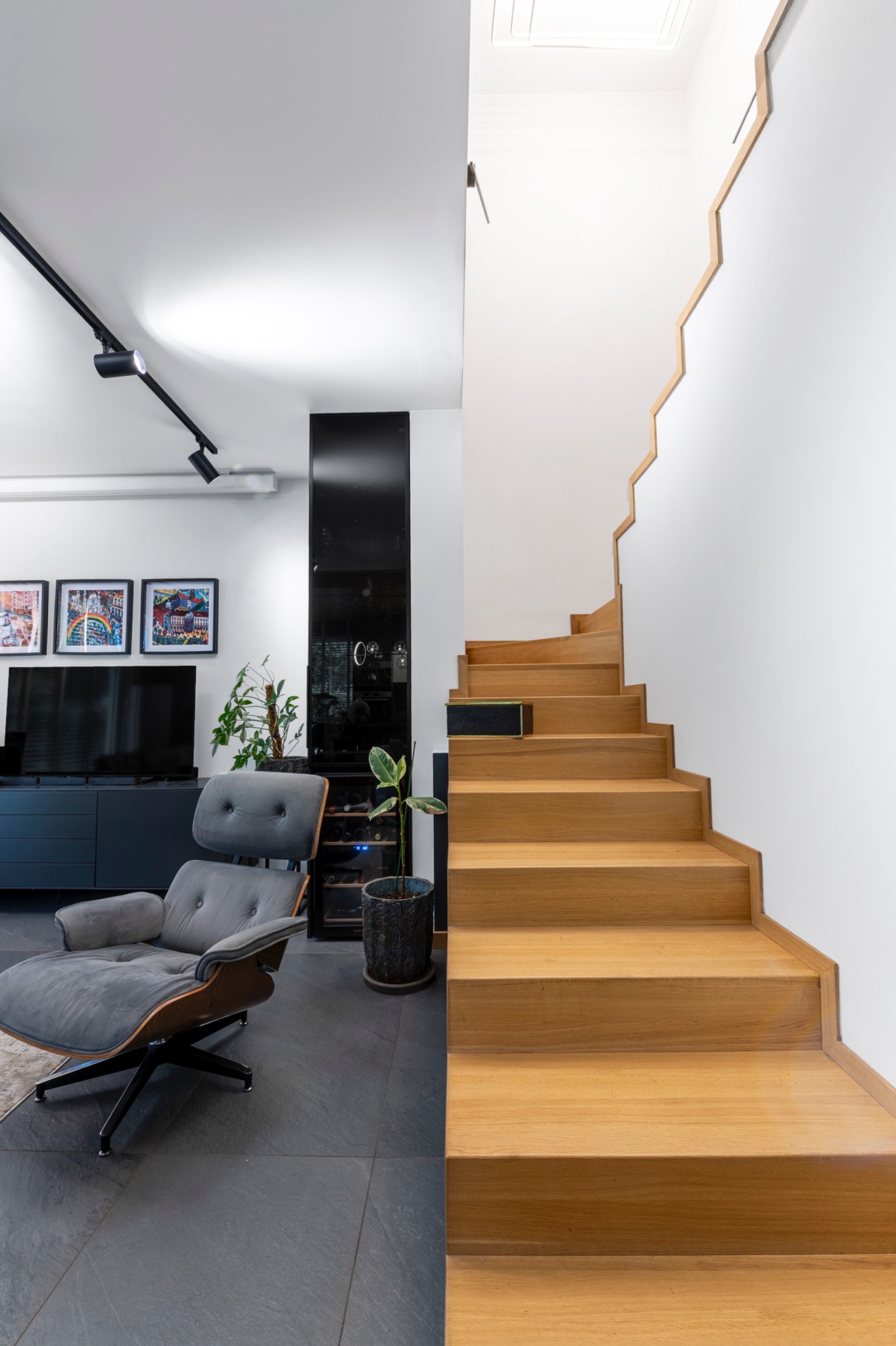 Nowoczesne wnętrze z drewnianymi schodami o nieregularnym kształcie, szarym fotelem i czarną zabudową z telewizorem i roślinami doniczkowymi.
