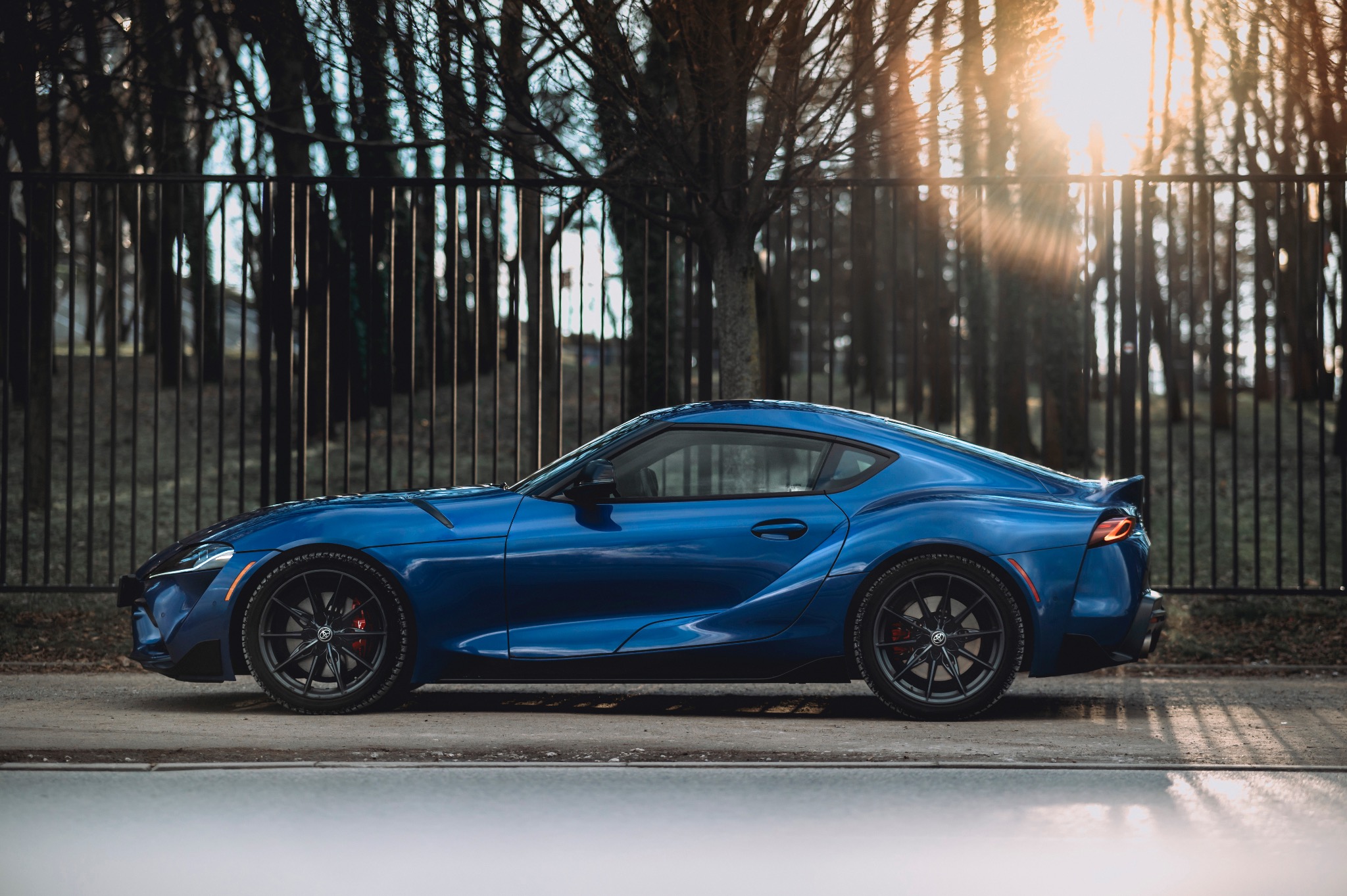 Niebieska Toyota Supra na tle metalowego ogrodzenia i drzew, z promieniami słońca prześwitującymi przez gałęzie.