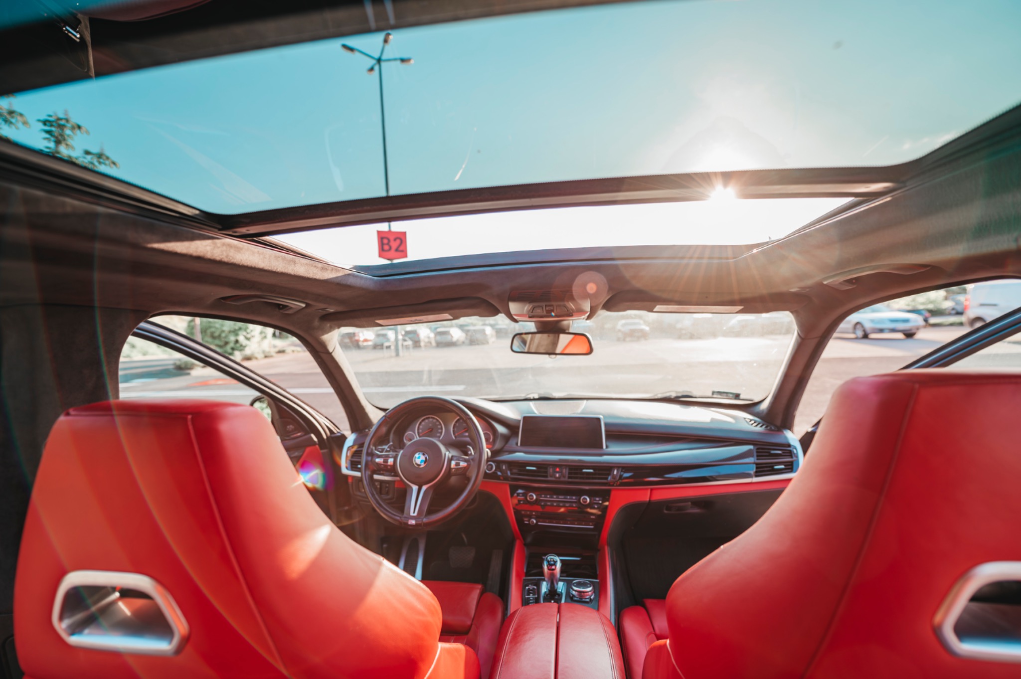 Wnętrze samochodu BMW X6 z czerwonymi skórzanymi fotelami i otwartym panoramicznym dachem, widok z tylnych siedzeń w kierunku deski rozdzielczej, z widocznym parkingiem na zewnątrz.