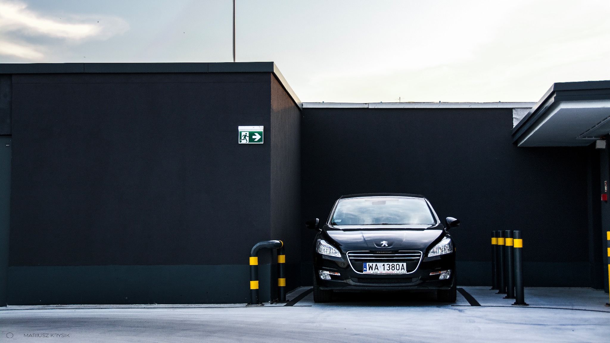 Czarny samochód Peugeot zaparkowany przy ciemnym budynku z oznakowaniem drogi ewakuacyjnej i słupkami ochronnymi z żółtymi pasami.