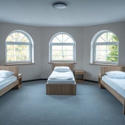 PRYWATNY OŚRODEK LECZENIA UZALEŻNIEŃ EXIT - Wnętrze pokoju z trzema łóżkami pojedynczymi, stoliki nocne, okna łukowe z widokiem na zieleń, szara wykładzina, neutralna kolorystyka ścian.