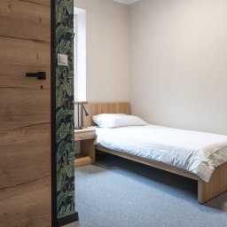 PRYWATNY OŚRODEK LECZENIA UZALEŻNIEŃ EXIT - Pokój z pojedynczym łóżkiem, drewnianą szafką nocną i lampką, widok zza drewnianych drzwi z czarną klamką i fragmentu ściany w ciemną tapetę z motywem liści