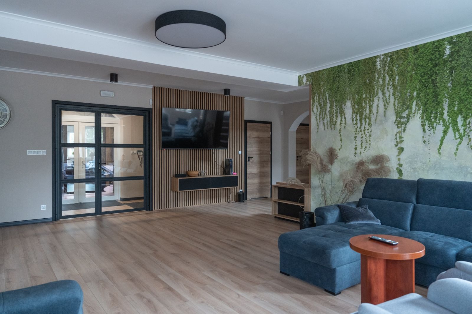 Nowoczesny salon z drewnianą ścianką TV, szklanymi drzwiami i ścianą z motywem pnączy, utrzymany w stonowanej kolorystyce.