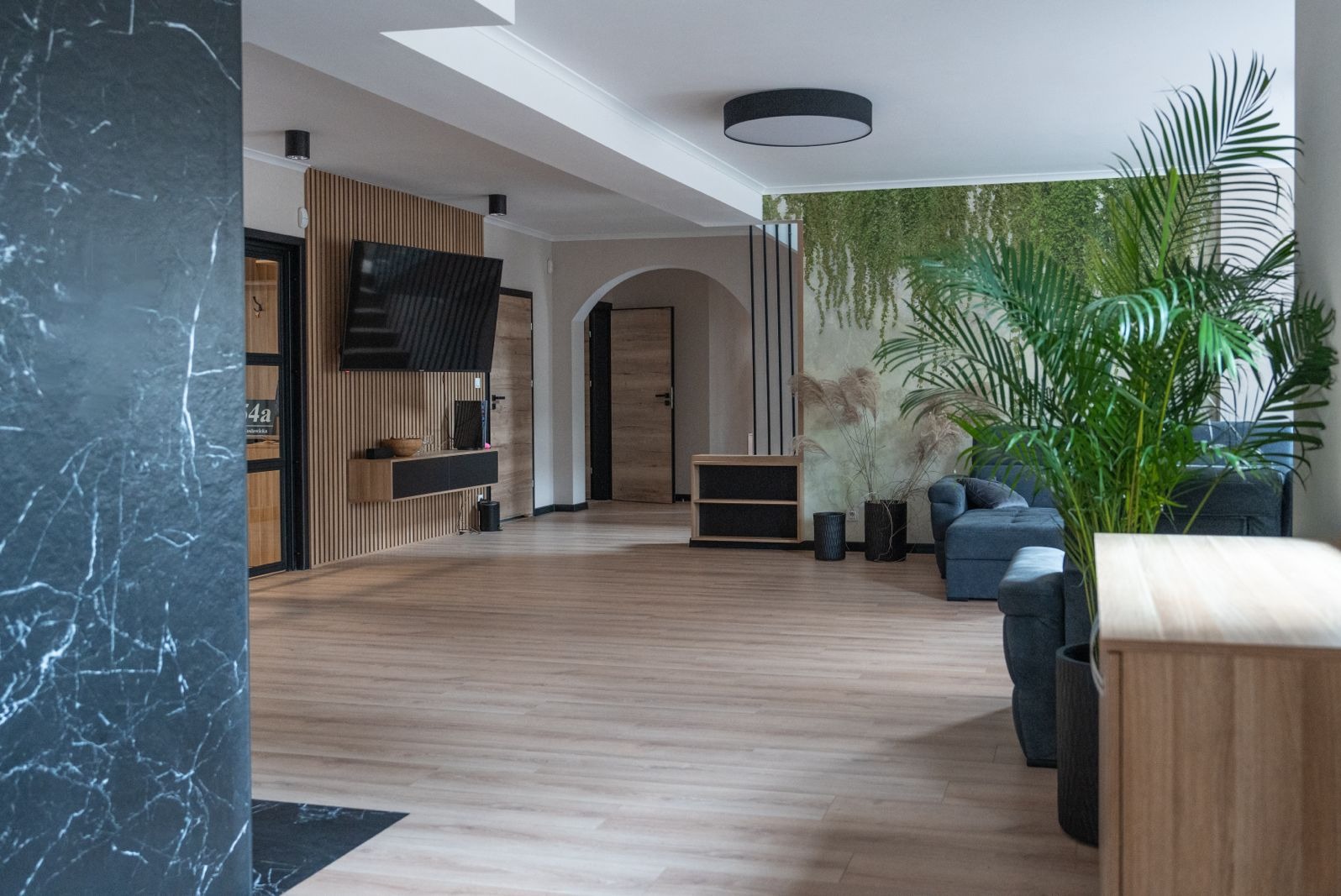 Jasne, przestronne wnętrze z elementami drewna, marmuru i zieleni, w tle kanapa i ściana z roślinnym motywem, nowoczesne oświetlenie sufitowe.
