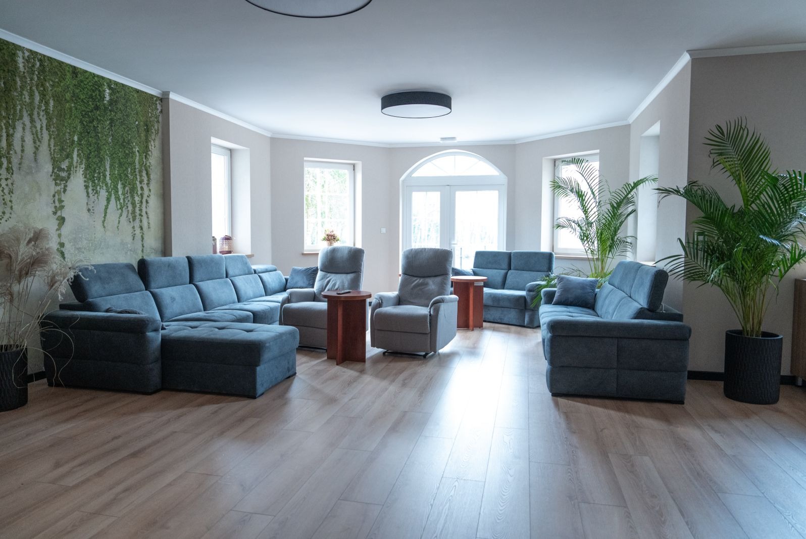 Przestronny pokój dzienny z szarymi sofami i fotelami, drewnianą podłogą i ścianą z motywem pnących roślin.