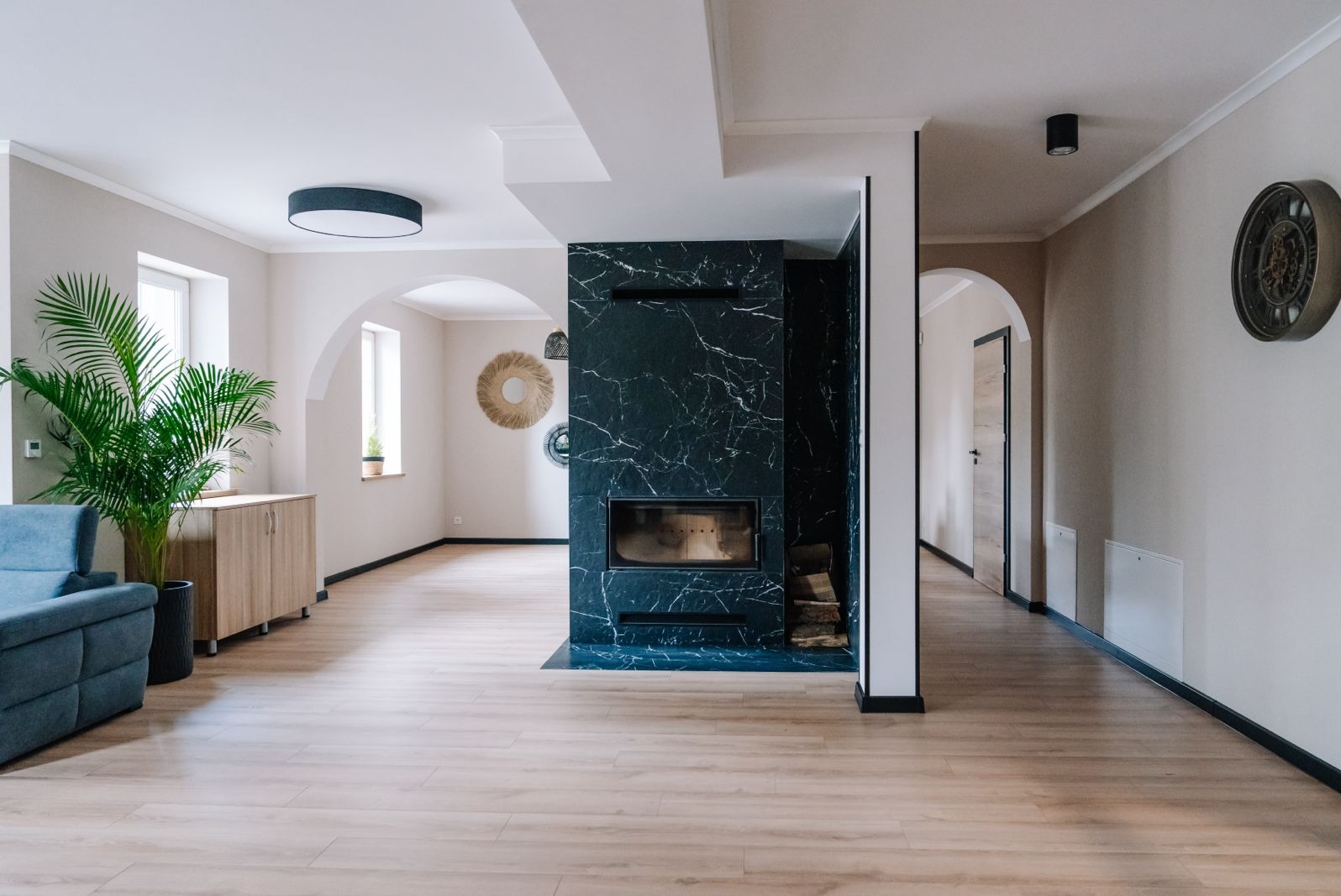 Przestronne wnętrze z kominkiem obłożonym czarnym marmurem z białymi żyłami, drewnianą podłogą i rośliną doniczkową w rogu pokoju, widoczne dekoracje ścienne i elementy architektoniczne jak łuki.
