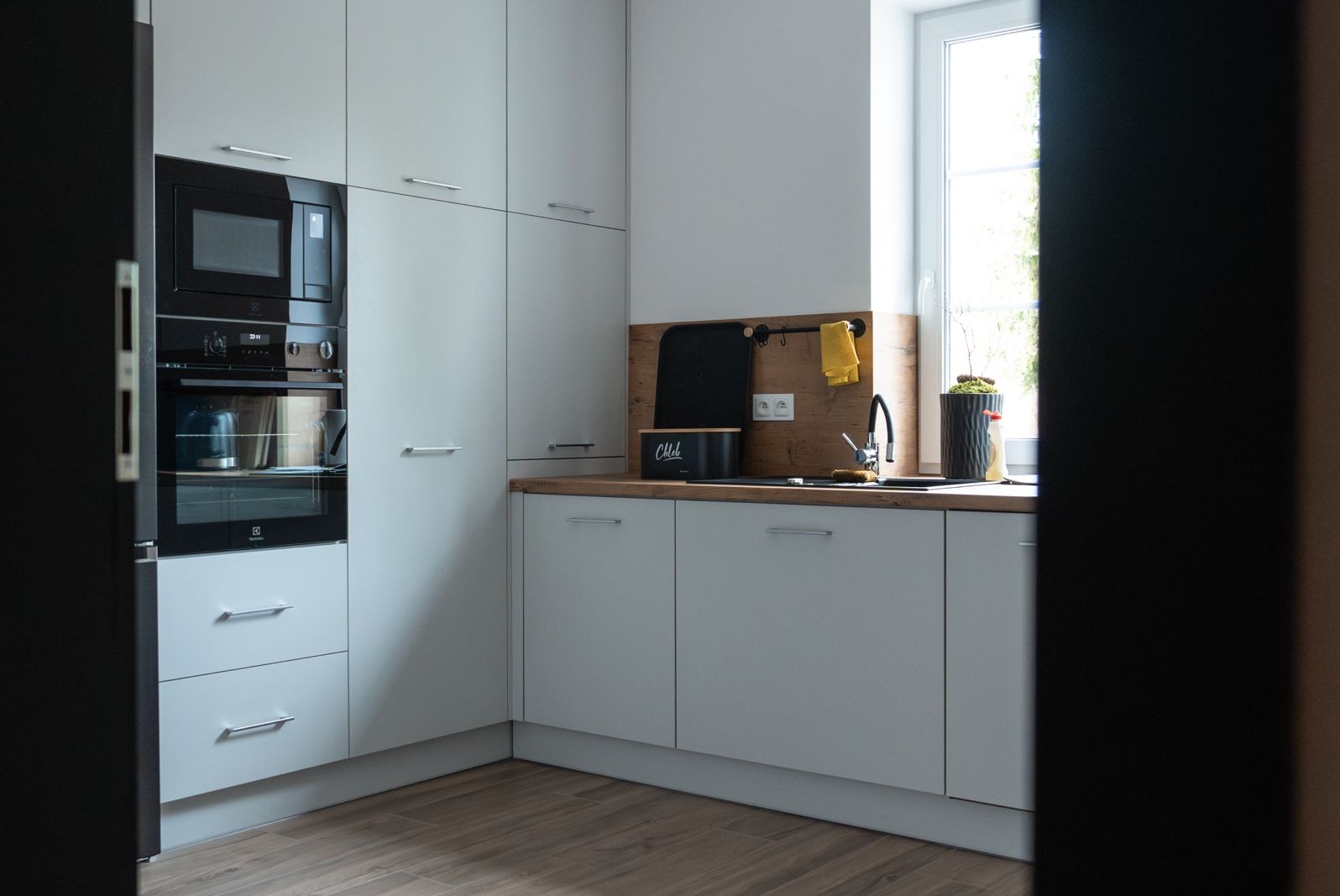 Jasna kuchnia z zabudową, piekarnikiem i mikrofalówką w czarnej obudowie, drewnianym blatem, czarnym zlewem i pojemnikiem na pieczywo z napisem 'Chleb'.