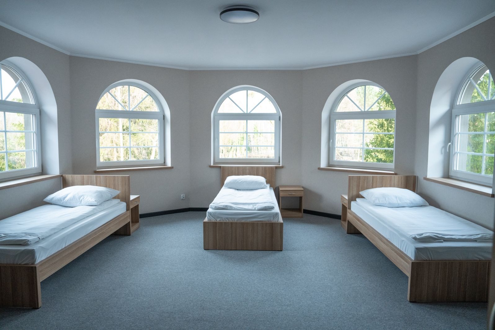 Wnętrze pokoju z trzema łóżkami pojedynczymi, stoliki nocne, okna łukowe z widokiem na zieleń, szara wykładzina, neutralna kolorystyka ścian.