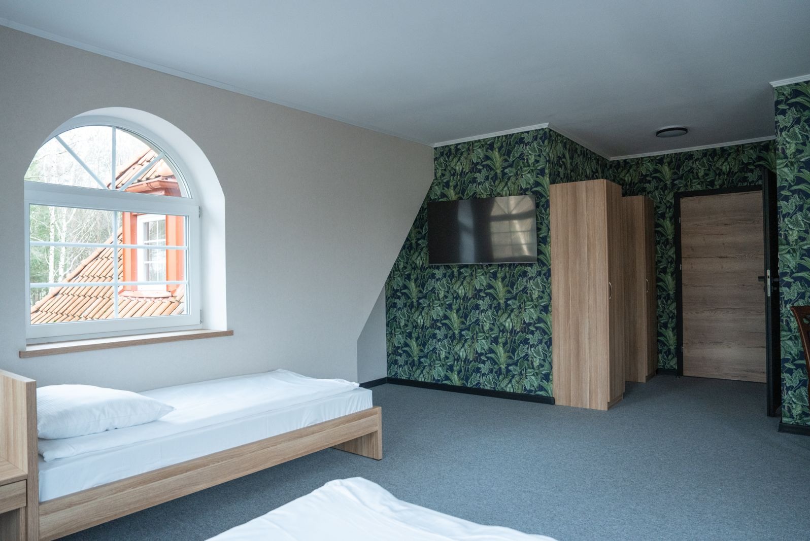 Pokój z dwoma łóżkami, oknem łukowym z widokiem na drzewa i ścianą w tropikalne liście z telewizorem.