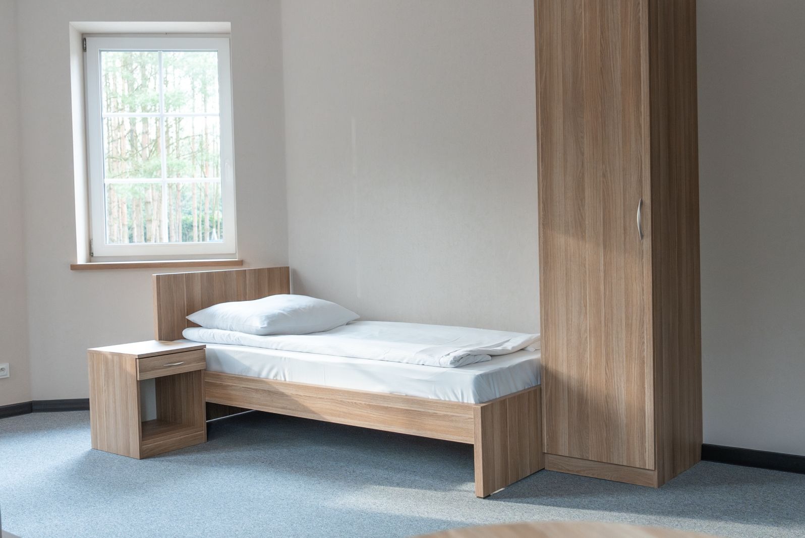 Pokój z pojedynczym łóżkiem, szafką nocną i szafą w odcieniach drewna, widok na las przez okno.