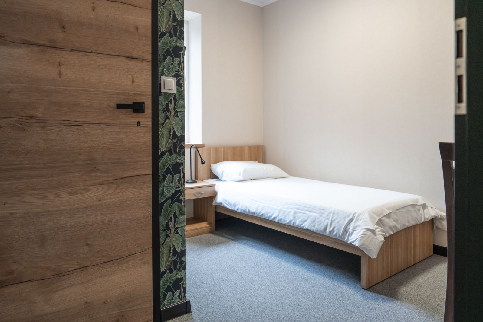 Pokój z pojedynczym łóżkiem, drewnianą szafką nocną i lampką, widok zza drewnianych drzwi z czarną klamką i fragmentu ściany w ciemną tapetę z motywem liści