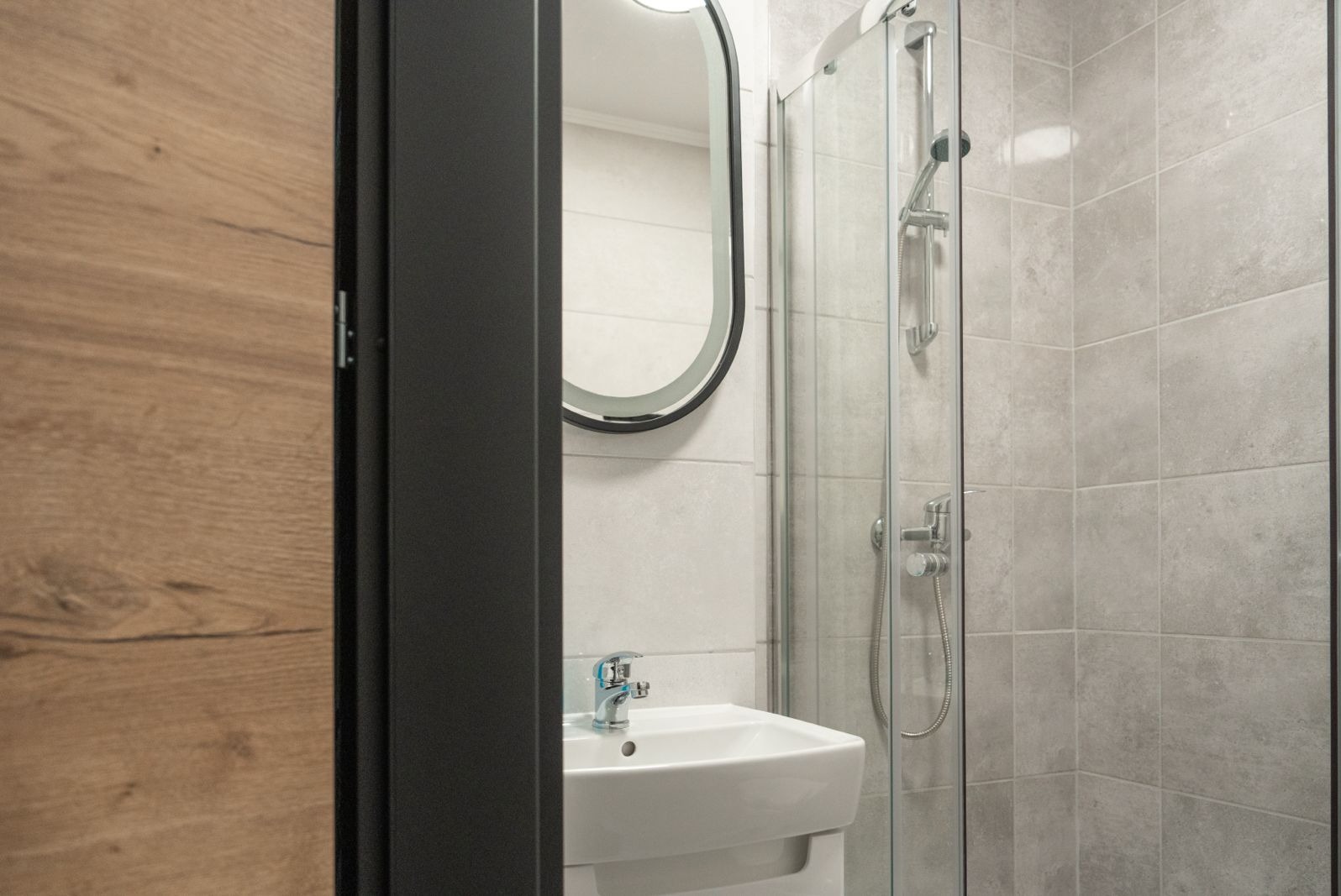 Nowoczesna łazienka z kabiną prysznicową, umywalką nablatową i owalnym lustrem z podświetleniem LED, widok zza uchylonych drzwi.